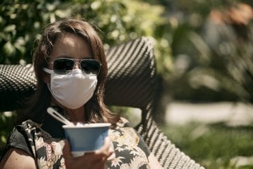 A woman holds a cup of ice cream while wearing a mask and sunglasses.