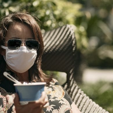 A woman holds a cup of ice cream while wearing a mask and sunglasses.