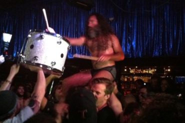 a man carries another man on his shoulders as a third man holds up a drum that the second man is beating