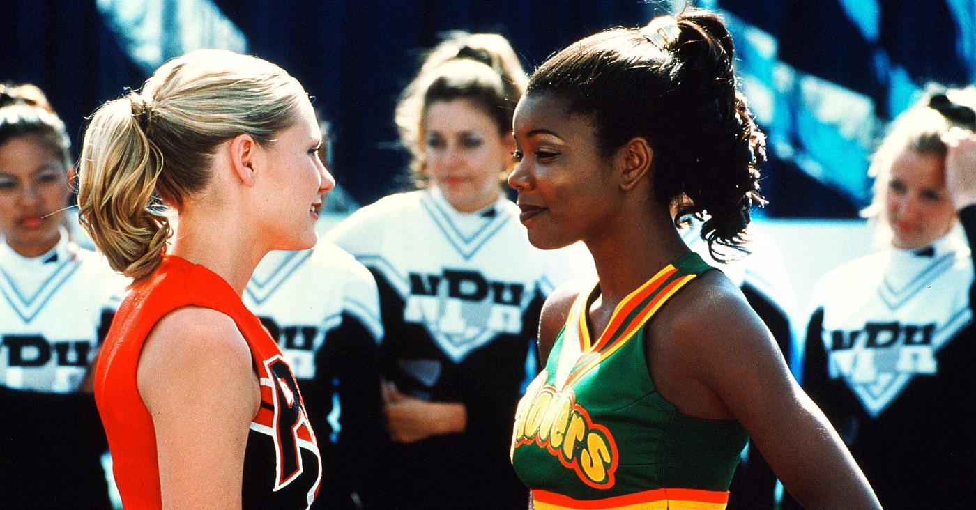 Two cheerleaders wearing rival team uniforms stare each other down while surrounded by other cheerleaders.