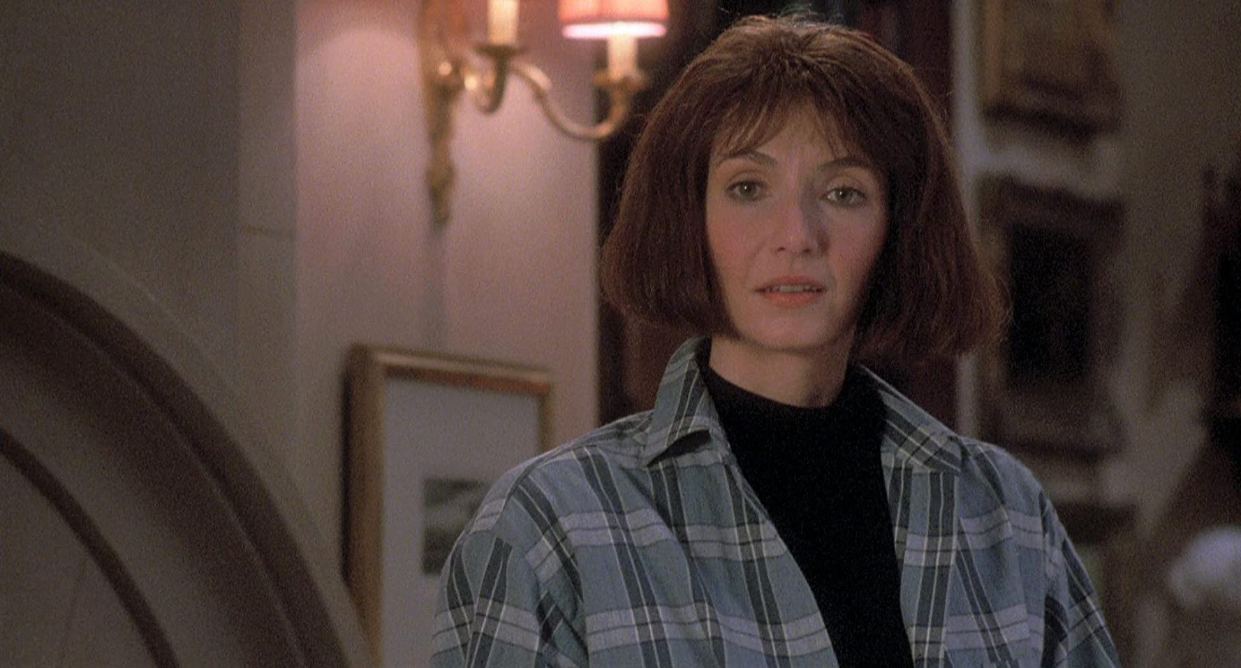 A woman wearing a plaid coat stands in a hallway decorated with picture frames, a look of concern on her face.