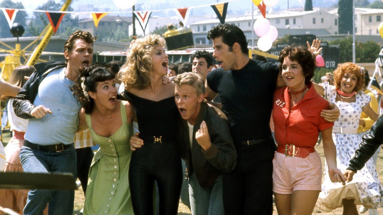 A group of 1950s teenagers stand together laughing and singing.