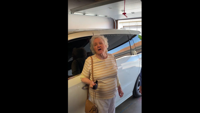 A video screenshot of an elderly woman standing in a garage and speaking to the person behind the camera.