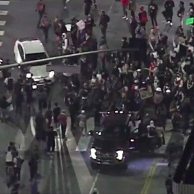 A nighttime overhead photo of an intersection crowded with people on foot. A white Prius is beginning to drive through the crowd.