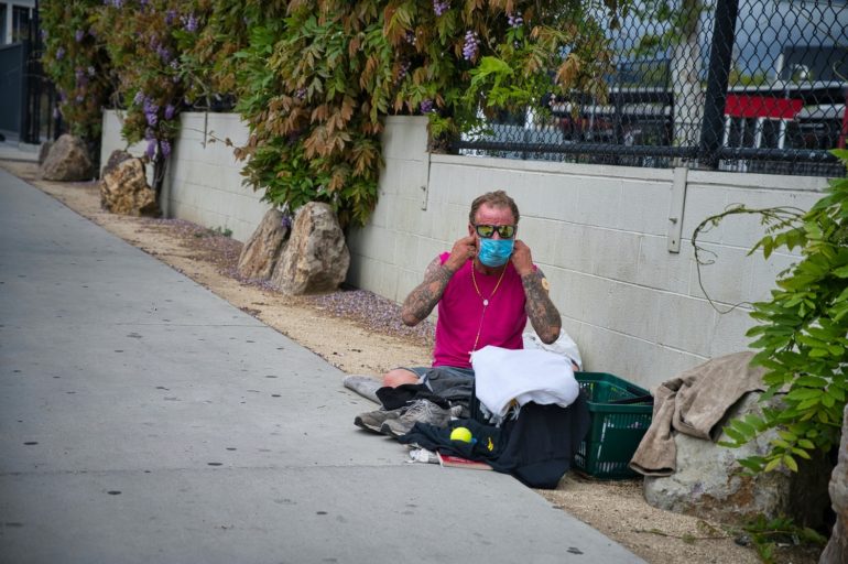A man in a pink shirt sits on a sidewalk next to his belongings.
