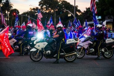 Police officers sit on motorcycles in the street, blocking off a crowd of pro-Trump demonstrators in the background who are holding Trump 2020 flags.