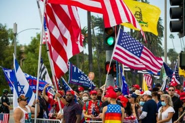 A crowd of people gathered on the sidewalk, waving United States flags, Trump 2020 flags, a "Don't tread on me" flag, and an Israel flag. Many also wear red "Make America Great Again" hats.