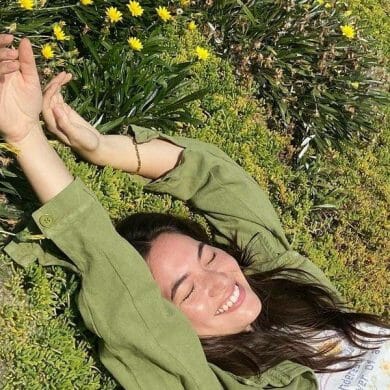 A model wearing a green jacket lays on green grass, smiling in the sunlight.